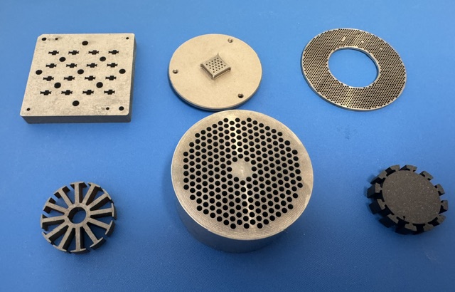 Assortment of precision-machined metal components on a blue background, including a perforated plate, a notched disc, a fine-hole ring, a finned wheel, a hexagonal-hole cylinder, and a small toothed gear